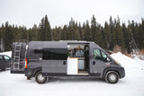 A grey Ram ProMaster rv rental, with open side and rear doors, parked in a snowy forest. Its custom interior is visible.
