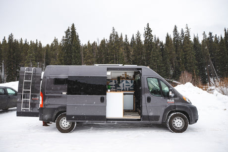 A grey Ram ProMaster rv rental, with open side and rear doors, parked in a snowy forest. Its custom interior is visible.