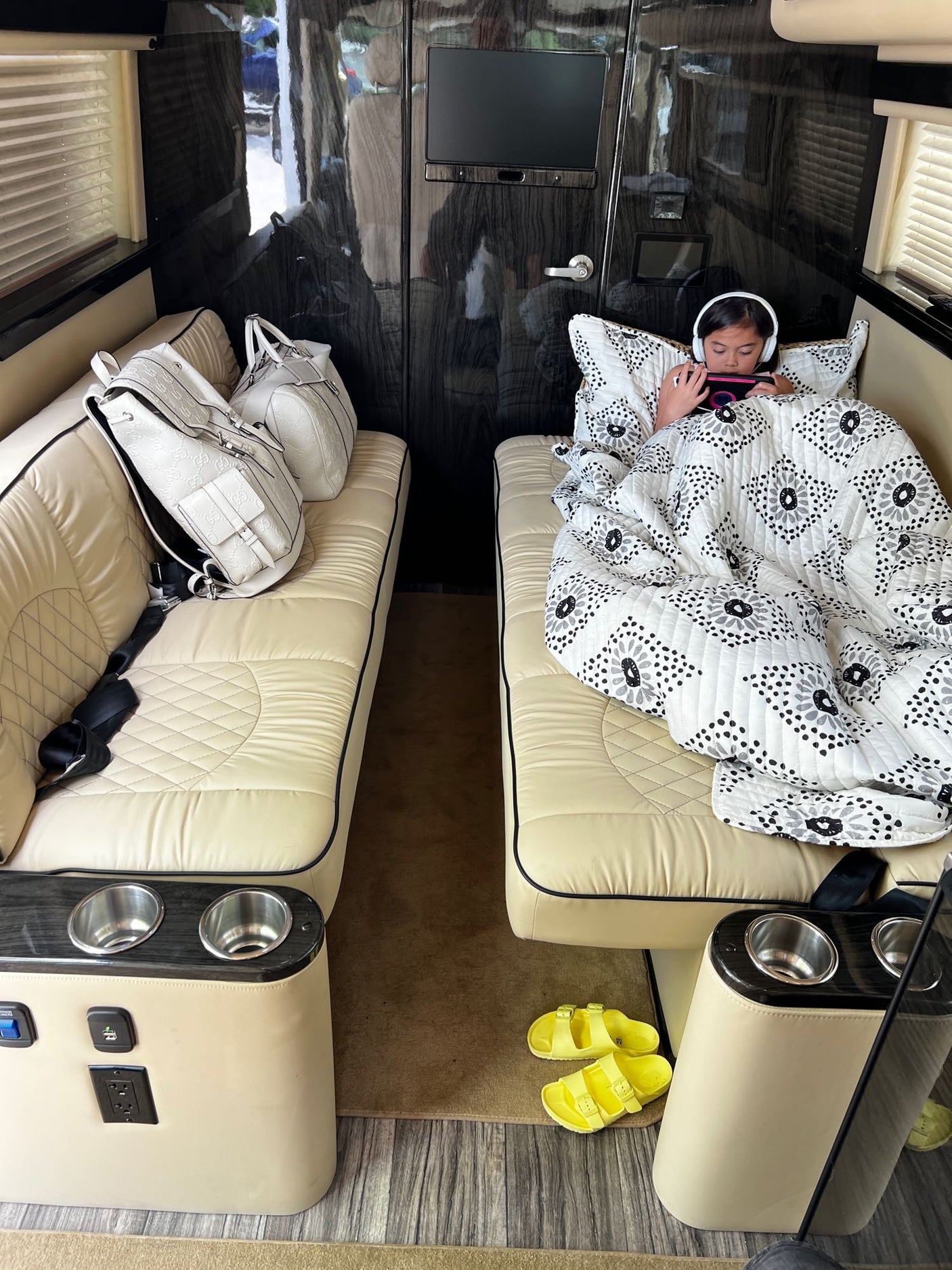 Relax in an **RV rental**! A child is cozy under a blanket on a plush bench with headphones, beside luxury bags.
