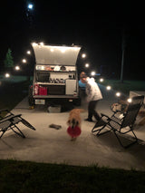 Nighttime RV rental setup with an open outdoor kitchen, string lights, a person, and a dog playing.