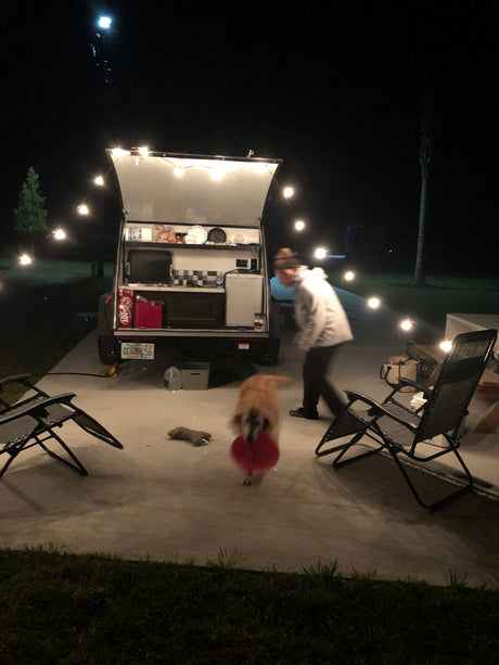 Nighttime RV rental setup with an open outdoor kitchen, string lights, a person, and a dog playing.