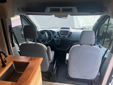 RV rental interior: View from rear of a camper van, showing a wooden kitchenette with sink, leading to the driver and passenger seats.