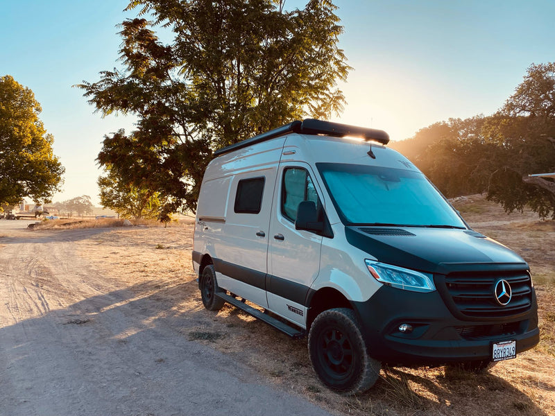  is leveled at a dirt campsite with doors open for adventure.|Rear view of a cream Winnebago Revel rv rental van with roof rack and ladder — photo 6