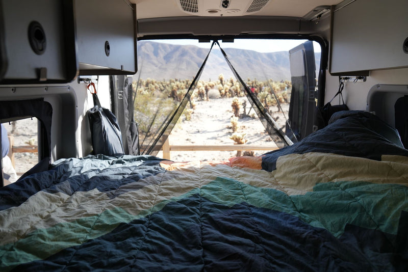 RV rental with a comfortable, patterned bed facing the open back, revealing a desert view of Cholla cacti and distant mountains. — photo 3