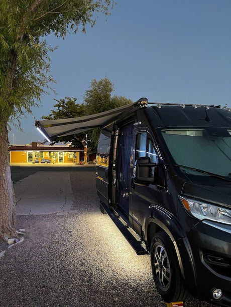 RV rental van at dusk with extended, lit awning, parked on gravel near a bright yellow building.