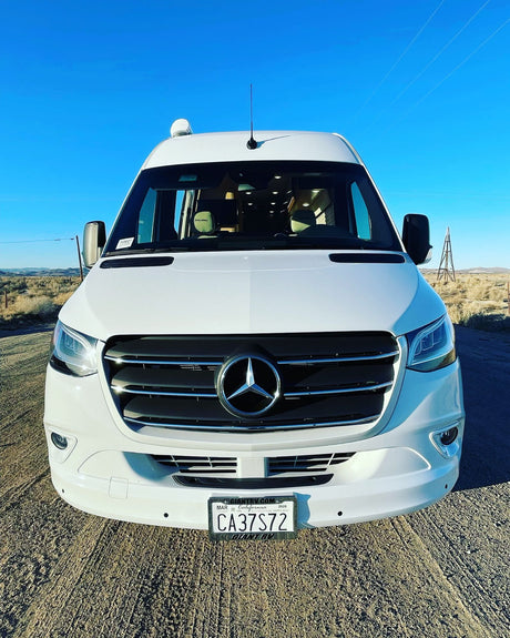 Front view of a white Mercedes-Benz Sprinter Galleria RV rental parked on a desert dirt road under a clear blue sky.