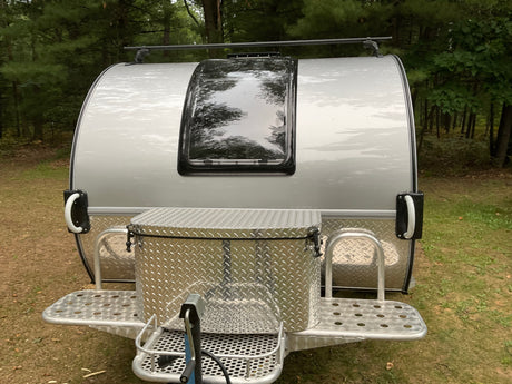 Silver teardrop travel trailer (RV rental) with a large front storage box and roof rack, parked in a wooded area.