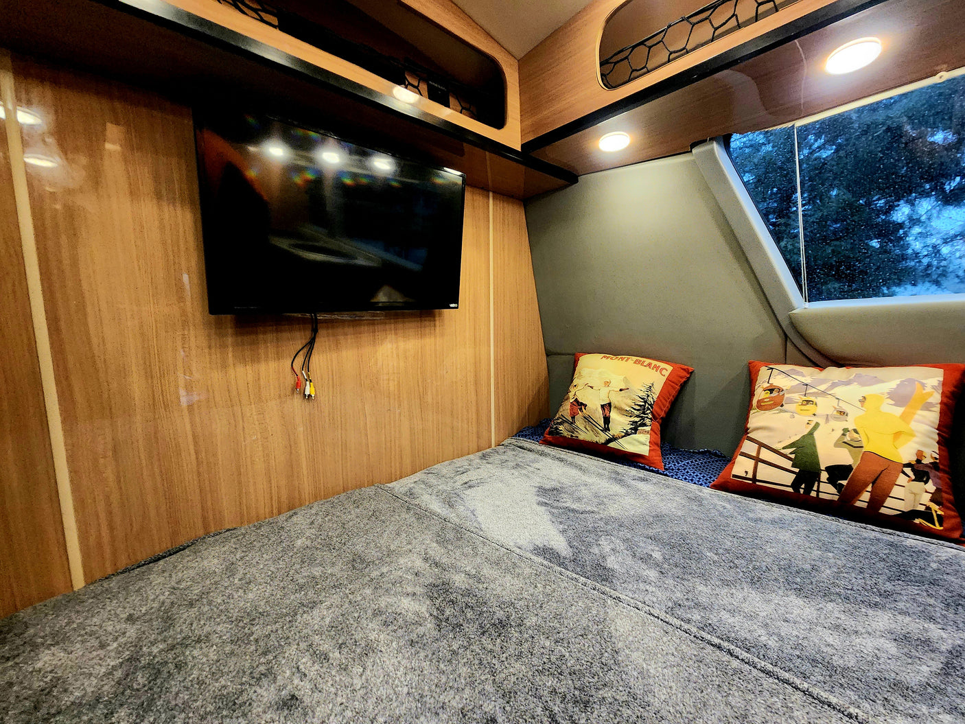 RV rental interior: cozy bedroom with a comfy bed, wall-mounted TV, and ski-themed pillows. A window shows trees outside.