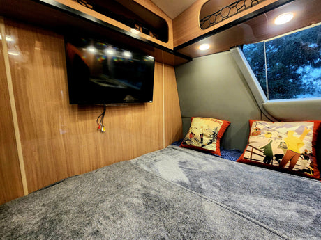 RV rental interior: cozy bedroom with a comfy bed, wall-mounted TV, and ski-themed pillows. A window shows trees outside.
