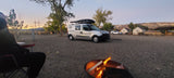An RV rental camper van at a gravel campground site, with a person by a glowing campfire under a sunset sky.