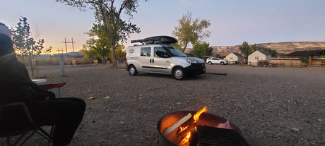 An RV rental camper van at a gravel campground site, with a person by a glowing campfire under a sunset sky.