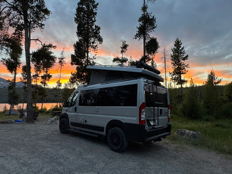 A grey RV rental camper van at a lakeside campsite, under a vibrant sunset. Ideal for camping trips.