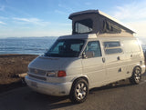 A white VW pop-top camper van by the ocean with mountains in the distance. Consider this RV rental for your next coastal trip!