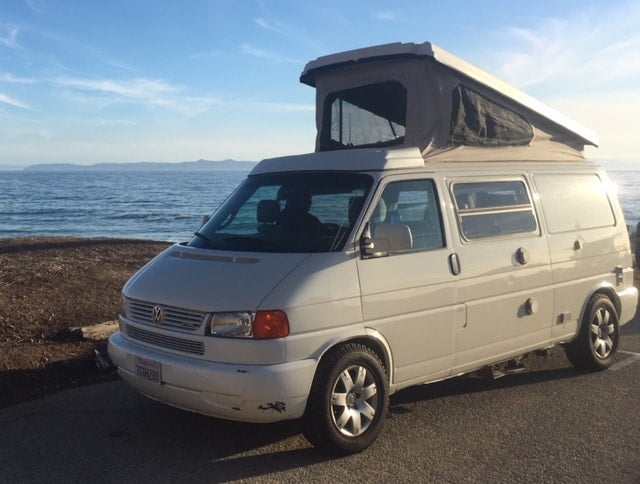  family plays UNO. A boy makes a funny face as kids enjoy cards with an ocean view.|Smiling man cooks breakfast on RV rental stove. Enjoy compact kitchen convenience for your travels!|Inside a compact RV rental — photo 9