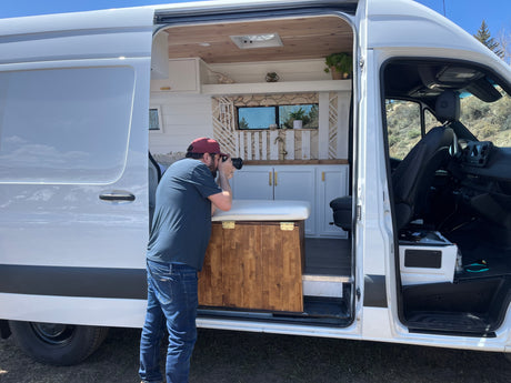 Man photographs the detailed, custom white camper van interior with wood ceiling, cabinets & sink. Perfect for an rv rental.