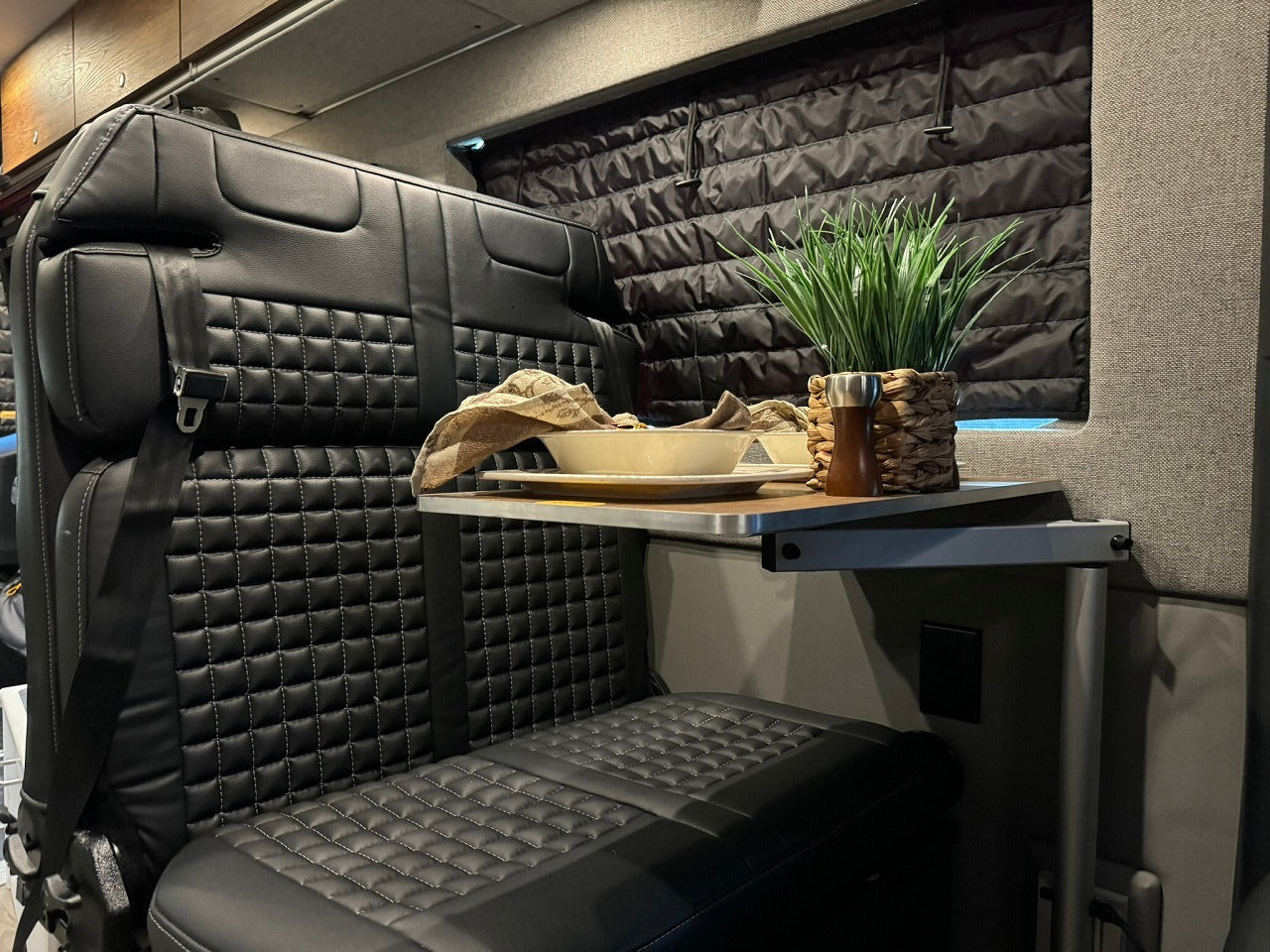 Cozy RV rental interior features quilted black seating, a folding table with a plant and meal setup, and insulated window cover.