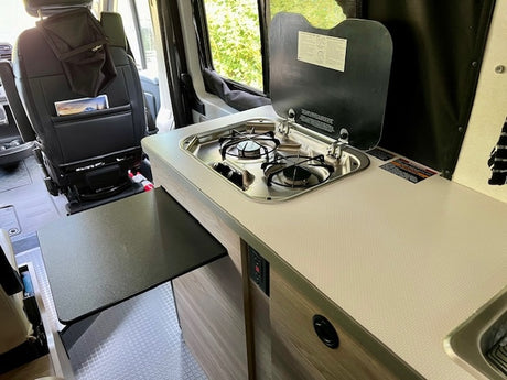 RV rental interior: compact kitchen with two-burner stove, sink, and pull-out table. Front seat storage visible for road trips.