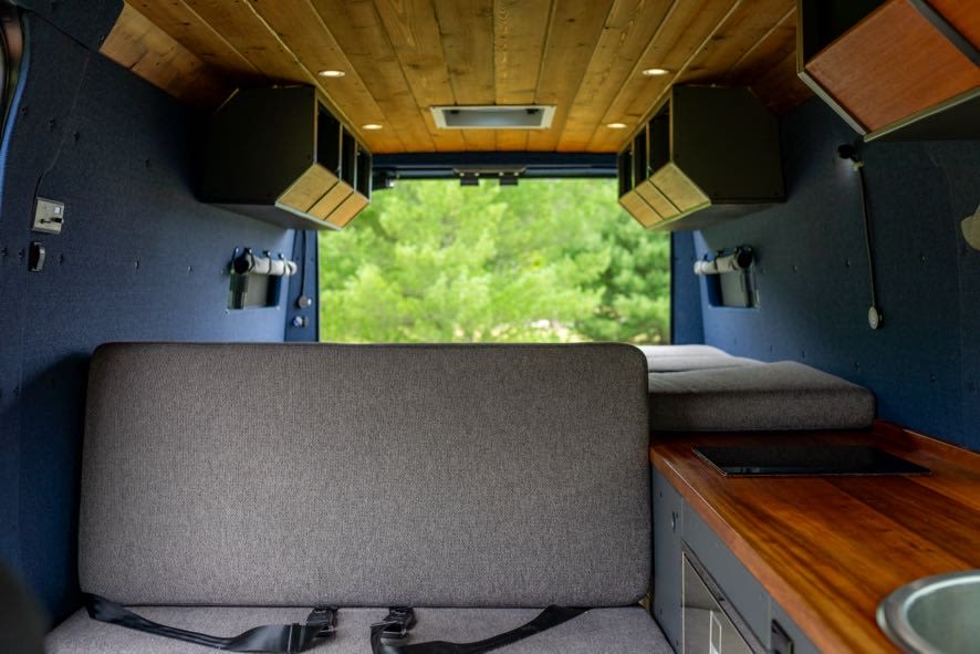 Inside a comfy RV rental camper van: bench seat/bed, wood kitchenette with sink/cooktop, overhead storage, and scenic rear view.