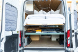Rear view of an **RV rental** van, doors open, revealing a neatly made bed, under-bed storage, and wood ceiling.