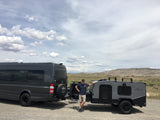 Man smiles by a dark grey RV rental van and compact trailer, ready for adventure in a vast, cloudy landscape.