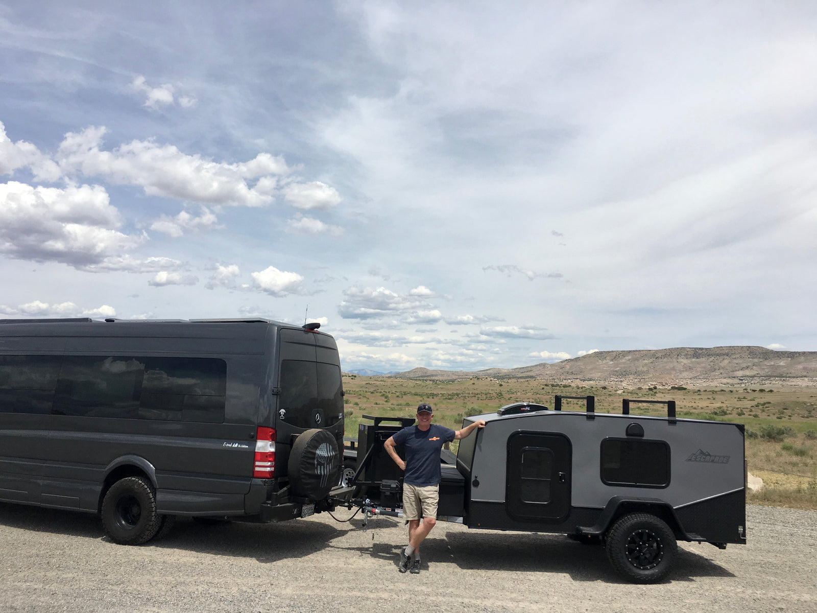 Man smiles by a dark grey RV rental van and compact trailer, ready for adventure in a vast, cloudy landscape.