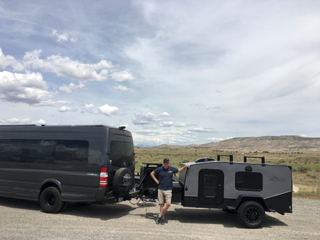 Man smiles by a dark grey RV rental van and compact trailer, ready for adventure in a vast, cloudy landscape.