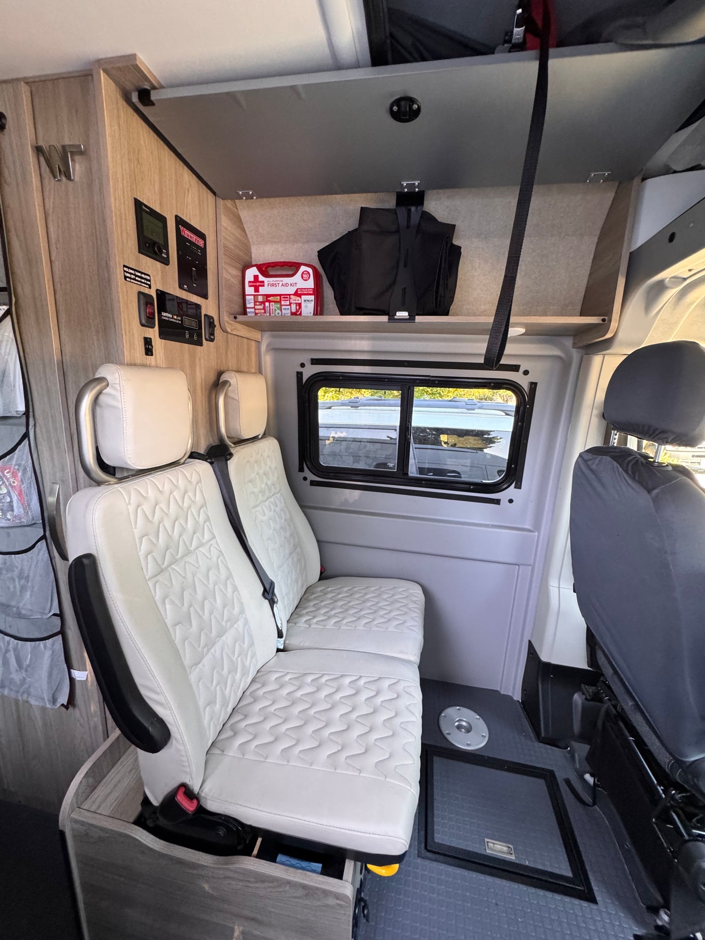 This camper van RV rental interior features white quilted passenger seats, essential control panels, overhead storage, and a window. — photo 11