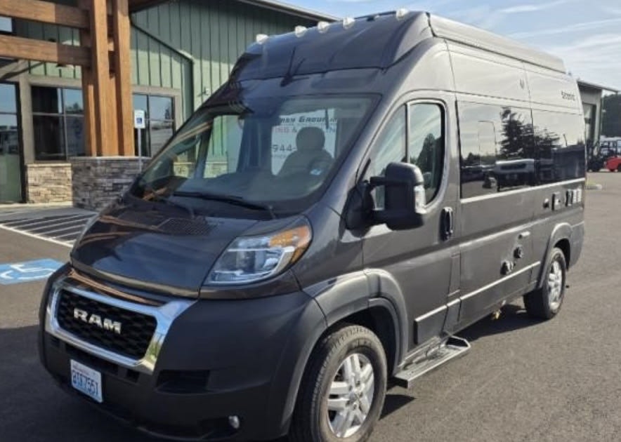 A dark grey Ram ProMaster high-roof campervan, ready for rv rental. It's parked outside a modern building.