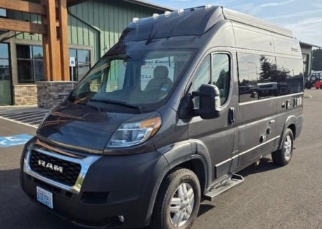 A dark grey Ram ProMaster high-roof campervan, ready for rv rental. It's parked outside a modern building.