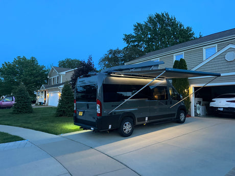 Grey RV rental with an illuminated awning extended in a suburban driveway at dusk.