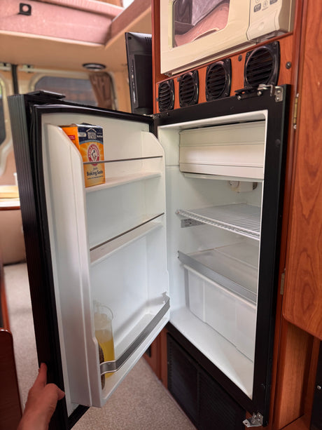 Inside an RV rental: open mini-fridge reveals baking soda and shelves. Compact kitchen setup with microwave.
