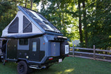 Rugged overland trailer with pop-up tent and solar panels, available for RV rental, parked in a scenic outdoor setting.