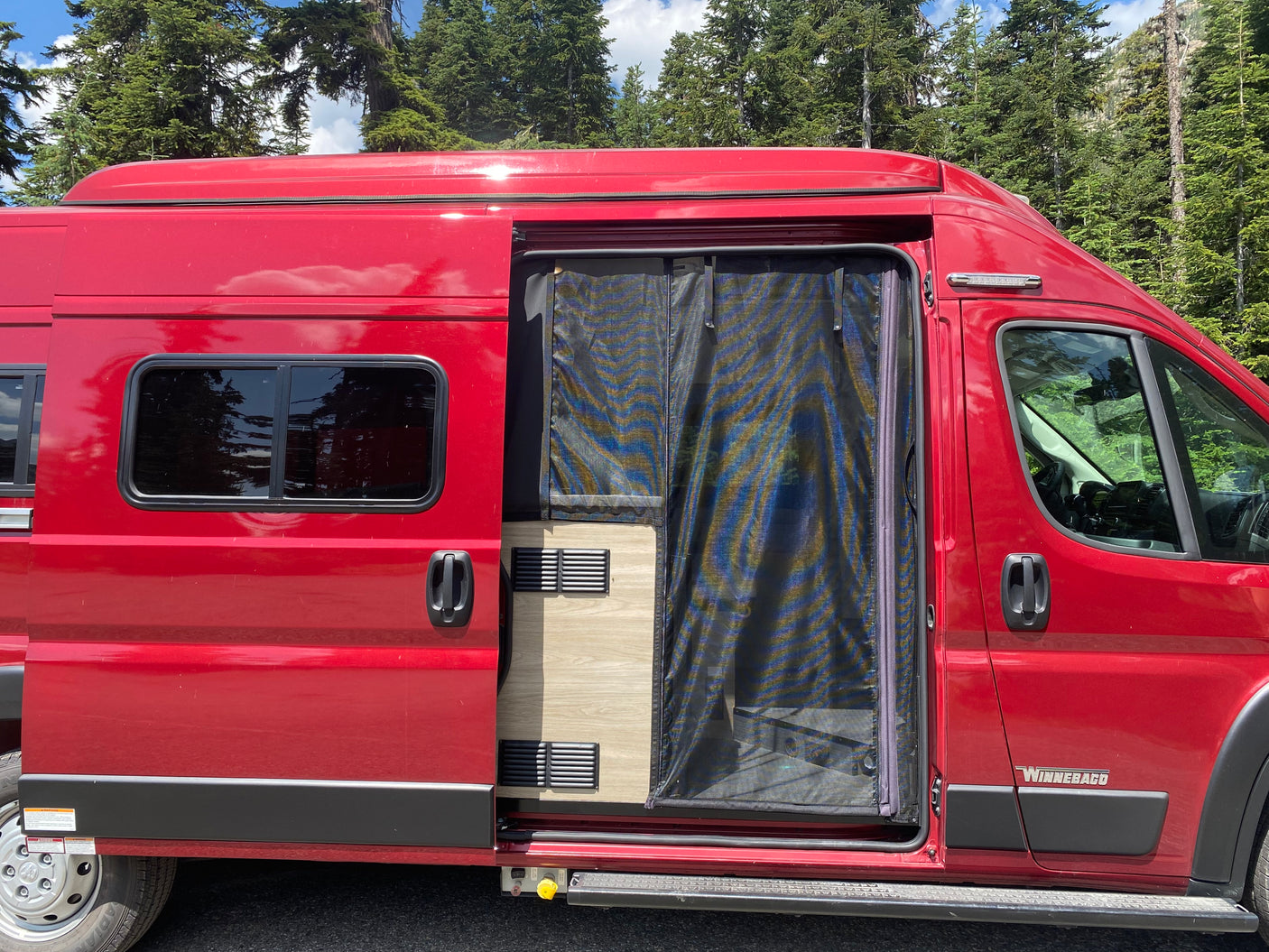 Red Winnebago camper van, an ideal RV rental, with open side door and screen. Parked by trees.
