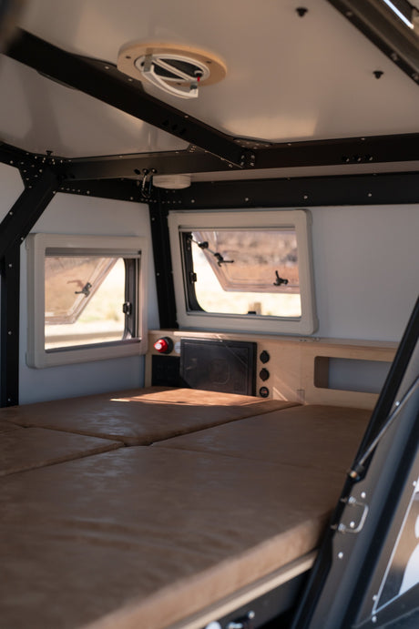 RV rental's compact interior: cushioned bed, open windows, ceiling fan, and built-in power panel.