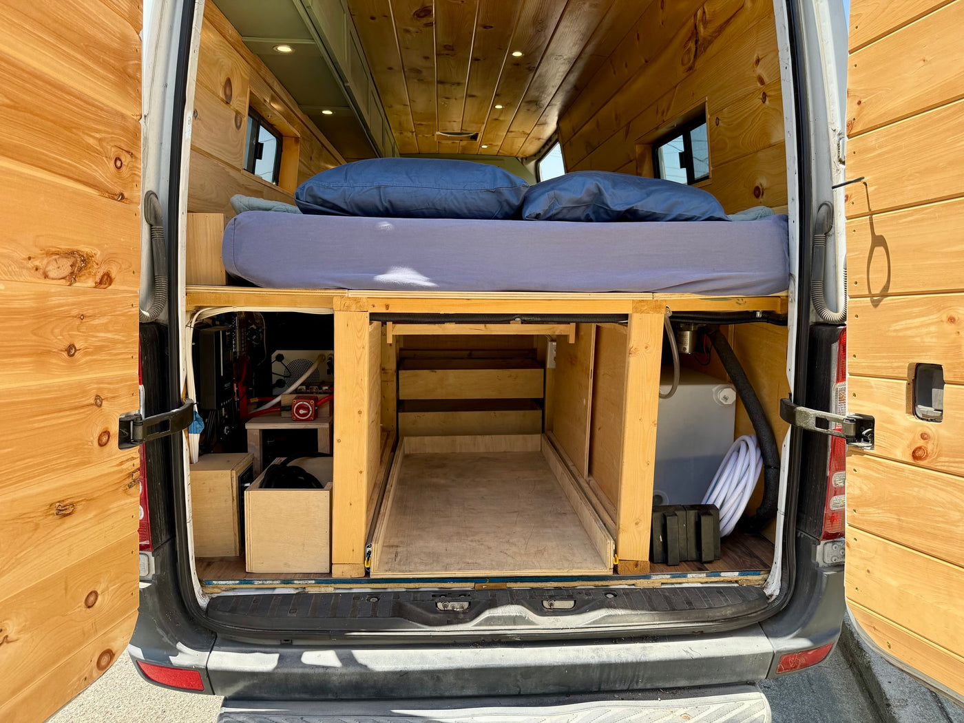View of a custom rv rental van's rear interior, showing a wooden bed platform, pull-out storage, and utilities. — photo 20