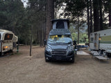 Modern grey RV rental campervan parked at a wooded campground between a pop-up and a travel trailer.