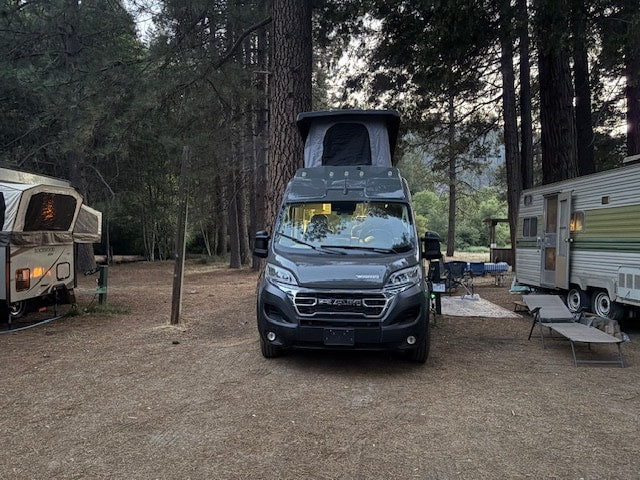  surrounded by tall pine trees and granite mountains.|Modern grey RV rental campervan parked at a wooded campground between a pop-up and a travel trailer.|Sleek gray Winnebago Solis campervan — photo 6