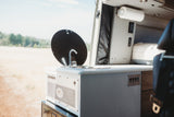 View inside an rv rental camper van: a compact kitchenette with sink, faucet, and controls. The rear door opens to a scenic outdoor field.