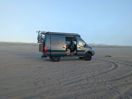 A dark teal camper van rv rental is parked on a sandy beach. Bikes are on the rear, a surfboard on the roof, and the side door is open.