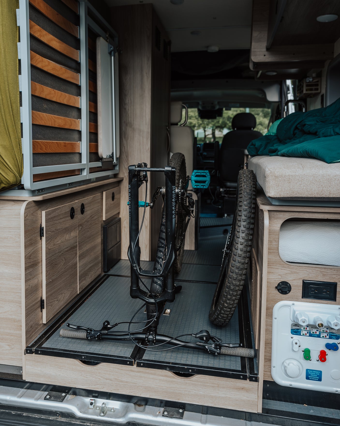 An RV rental interior with a mountain bike neatly stored for transport, featuring custom cabinetry, bed, and utility connections.