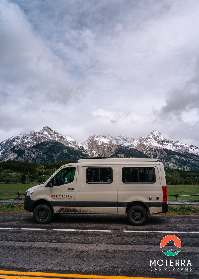 Beige Moterra RV rental campervan on a road, with majestic snow-capped mountains and green forests in the background.
