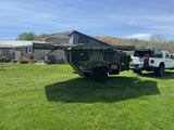 Off-road RV rental: a green overland trailer with an awning extended, hitched to a white Jeep Gladiator, ready for adventure.