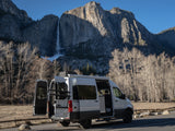 White RV rental camper van parked with open doors, overlooking Yosemite Falls and imposing mountain cliffs under a clear sky.
