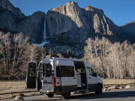 White RV rental camper van parked with open doors, overlooking Yosemite Falls and imposing mountain cliffs under a clear sky.