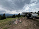Scenic RV rental campsite: Teardrop trailer with bikes, a fire pit, and mountain views under a cloudy sky.