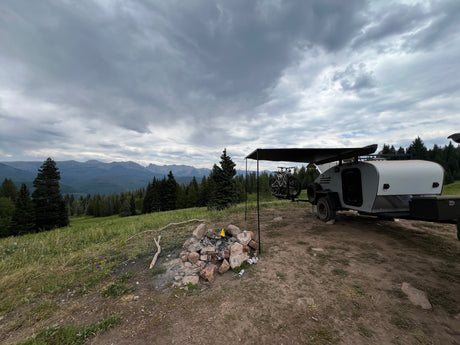 Scenic RV rental campsite: Teardrop trailer with bikes, a fire pit, and mountain views under a cloudy sky.
