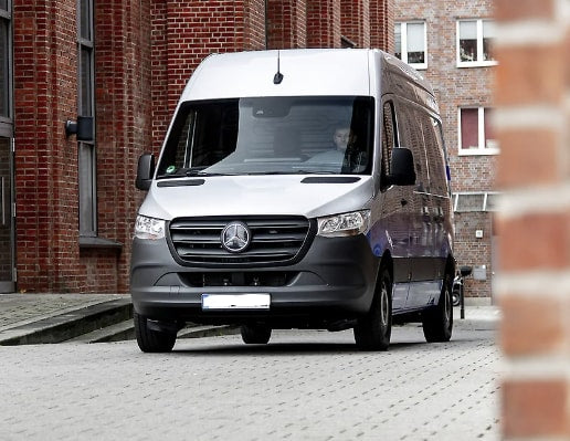 A silver Mercedes-Benz Sprinter van, a versatile RV rental, on a paved surface by brick buildings. Driver visible.