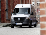 A silver Mercedes-Benz Sprinter van, a versatile RV rental, on a paved surface by brick buildings. Driver visible.