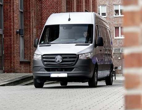 A silver Mercedes-Benz Sprinter van, a versatile RV rental, on a paved surface by brick buildings. Driver visible.