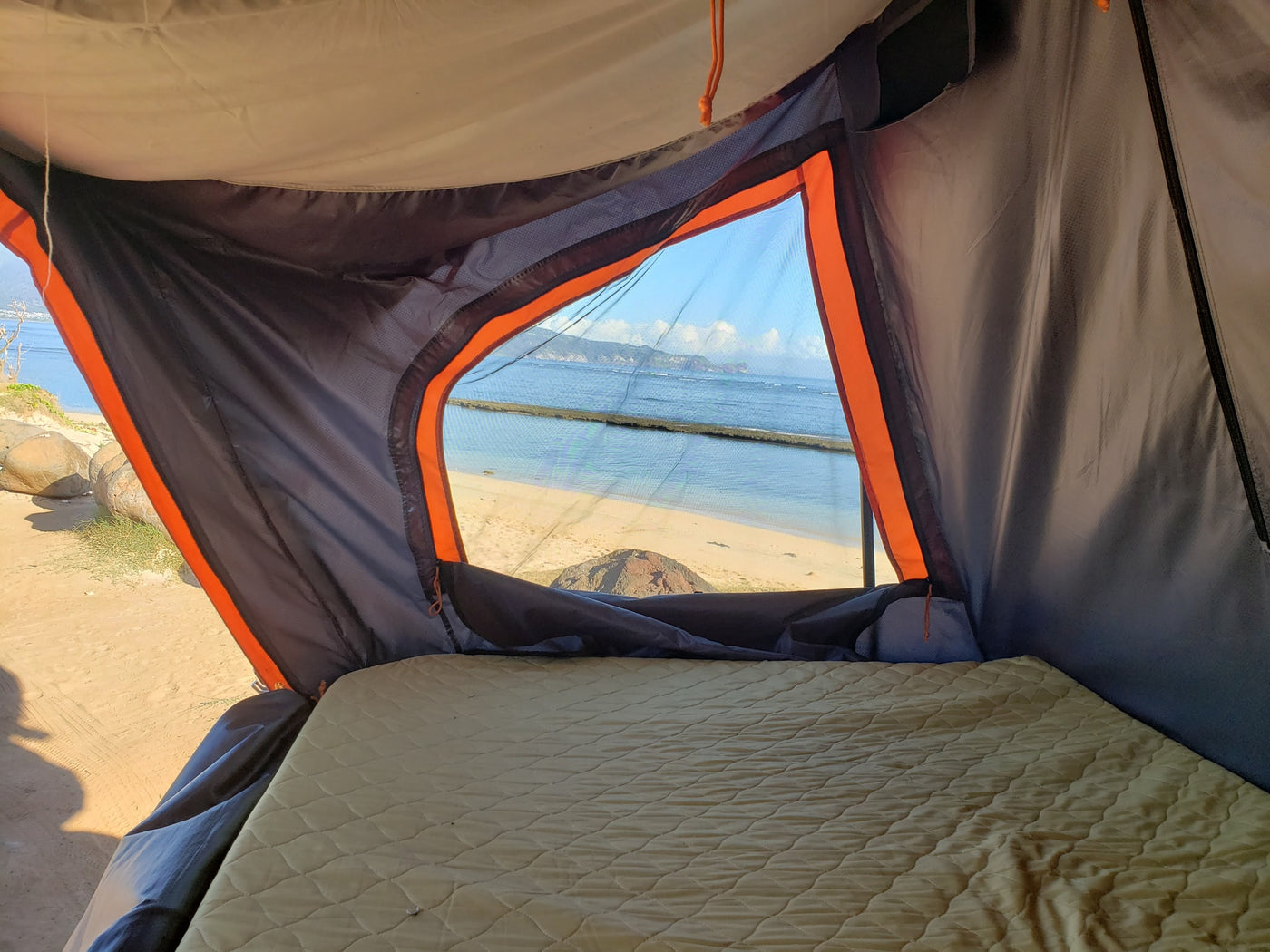 Inside an RV rental tent, a comfy mattress faces a screened window overlooking a serene beach and ocean view with distant mountains. — photo 2